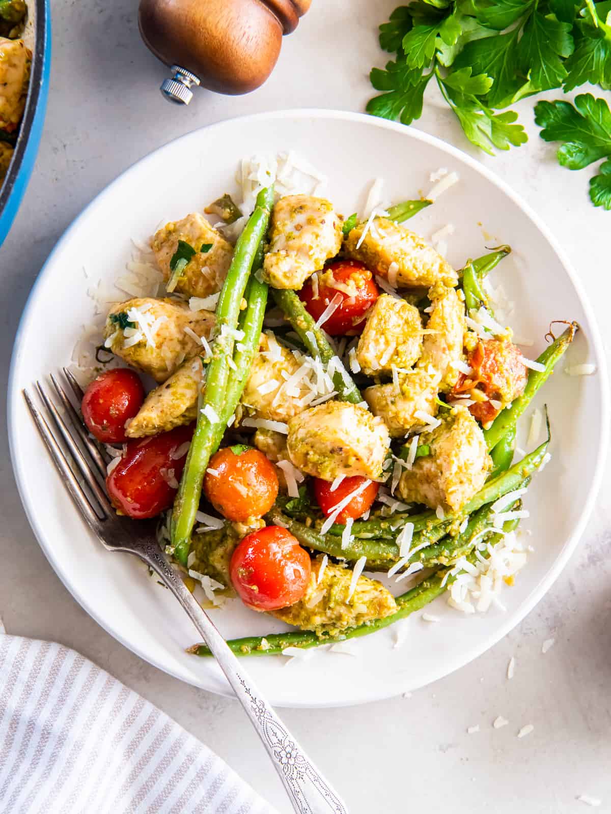 A serving of pesto chicken with green beans and tomatoes topped with Parmesan on a white plate.