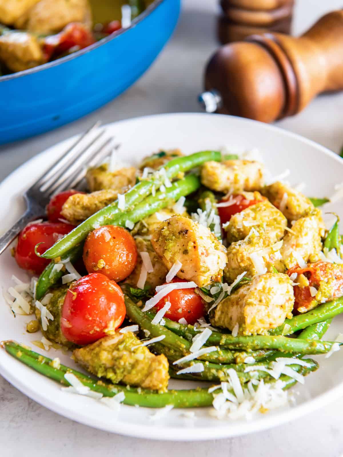 A serving of pesto chicken with green beans and tomatoes topped with Parmesan on a white plate in front of a skillet.