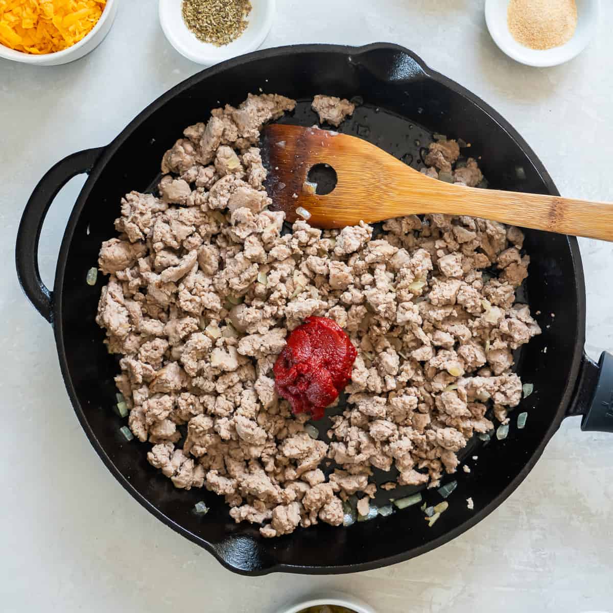 Tomato paste on top of cooked ground turkey in a cast iron skillet with a wooden spatula.
