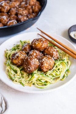 Asian meatballs over zucchini noodles on a white plate with chopsticks lying next to it.