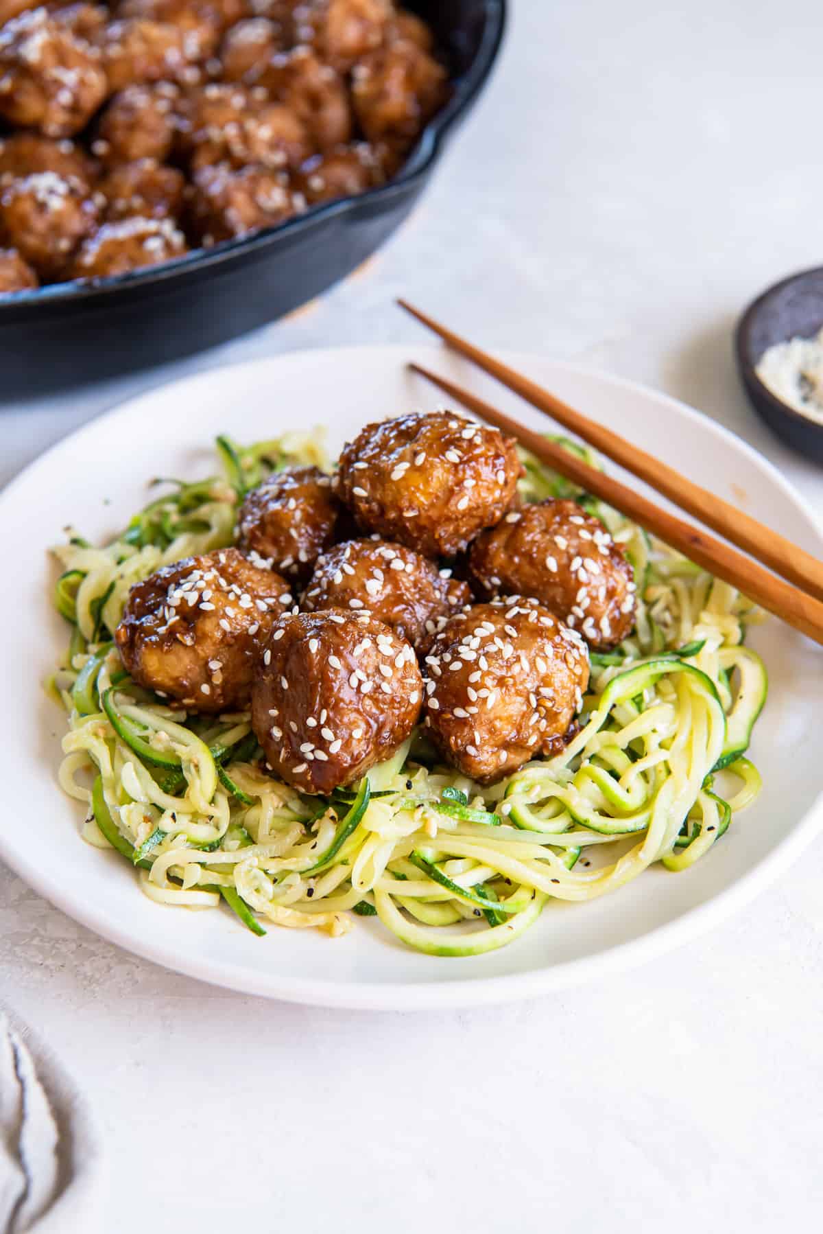 Asian meatballs over zucchini noodles on a white plate with chopsticks lying next to it.