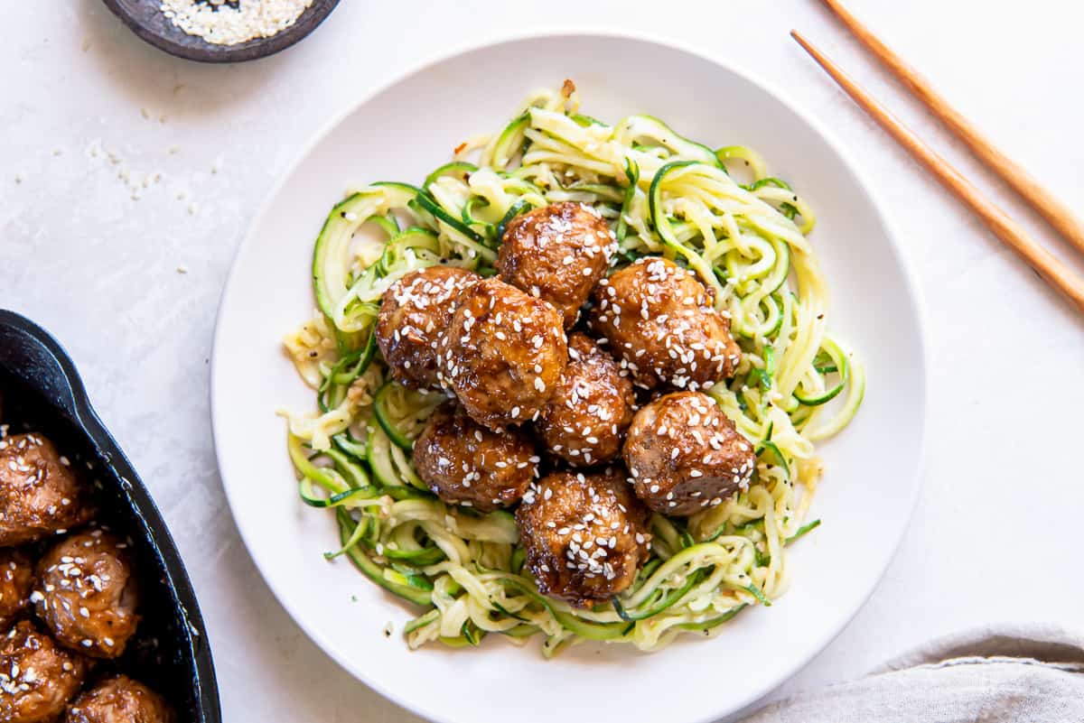 A top down shot of Asian meatballs over zucchini noodles on a white plate with chopsticks lying next to it.