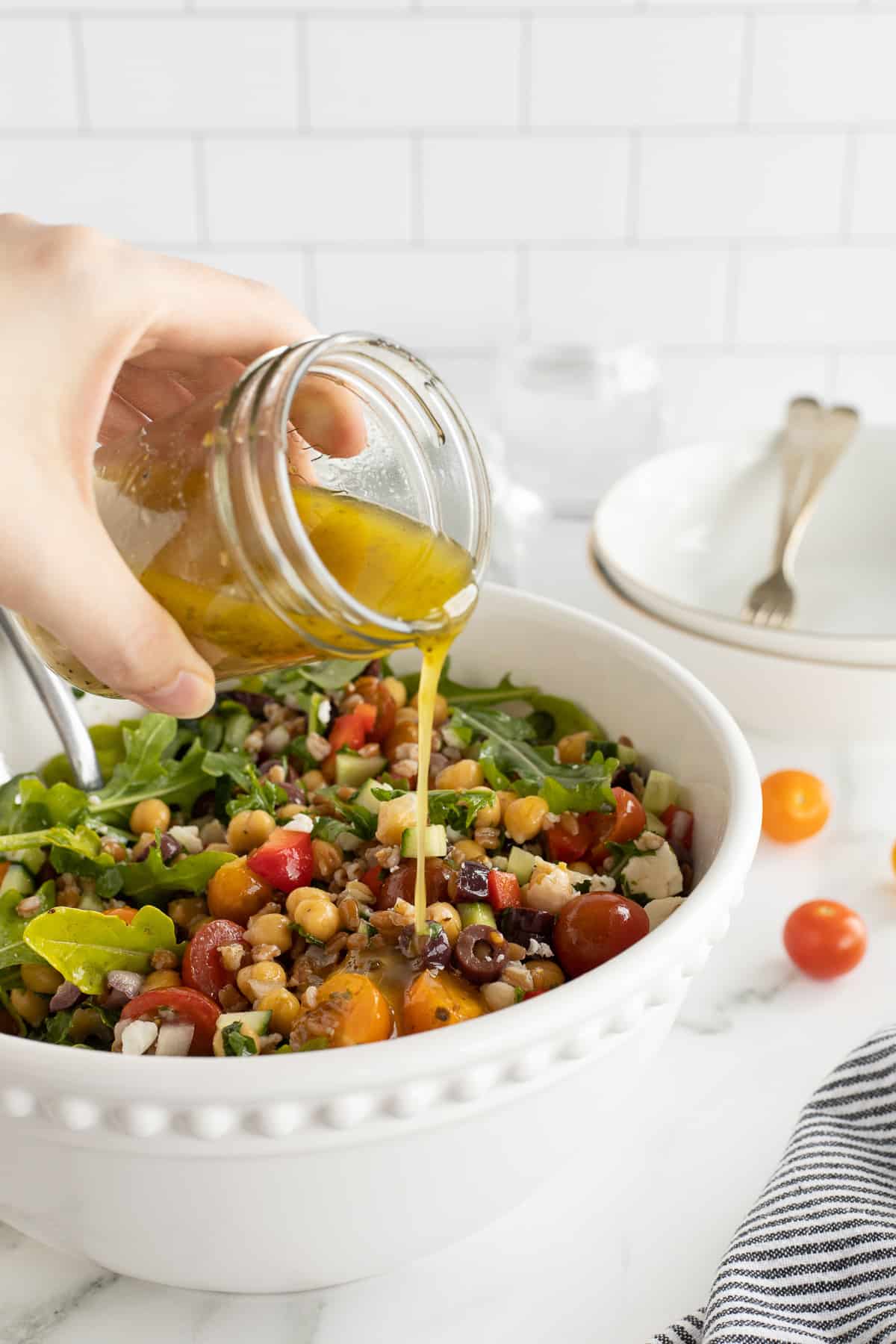 A hand pouring Greek salad dressing from a mason jar over a salad in a large white bowl.