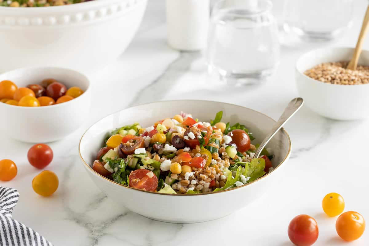 Greek farro salad in a white salad bowl with a fork with cherry tomatoes scattered around it.