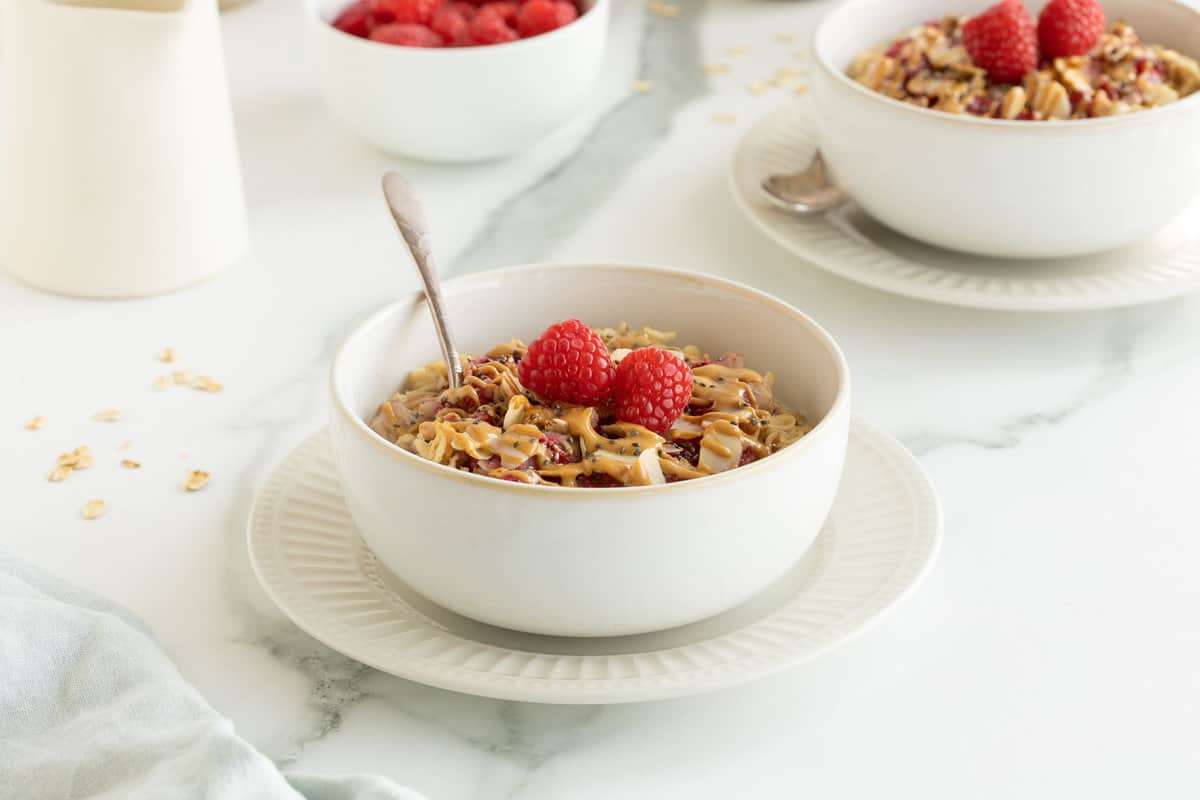 A side view of two bowls of oatmeal topped with raspberries and peanut butter on a kitchen counter.