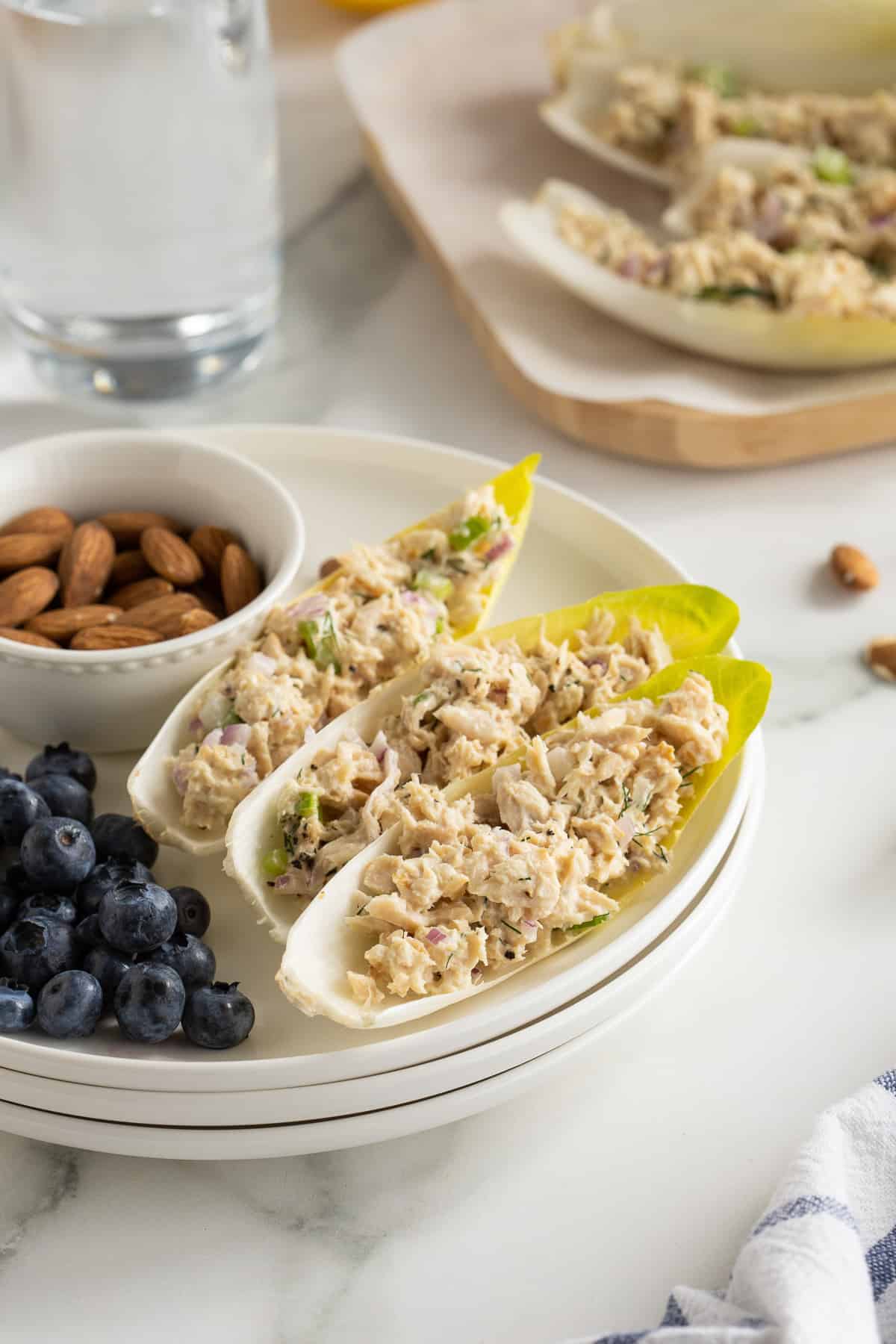 A side view of endive leaves stuffed with tuna salad on a white plate with blueberries and a small bowl of almonds.
