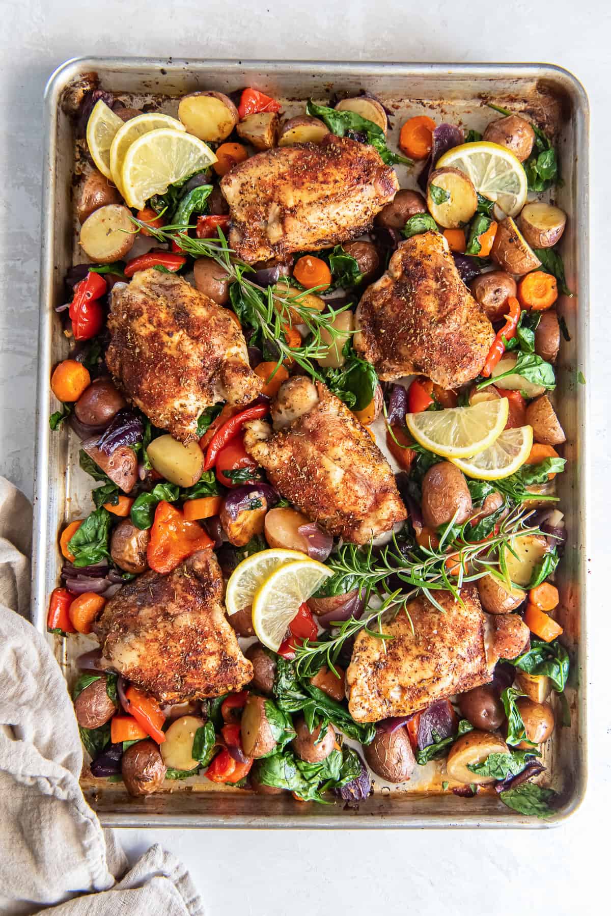 A top down shot of roasted chicken and vegetables on a sheet pan topped with lemon slices and fresh herbs.