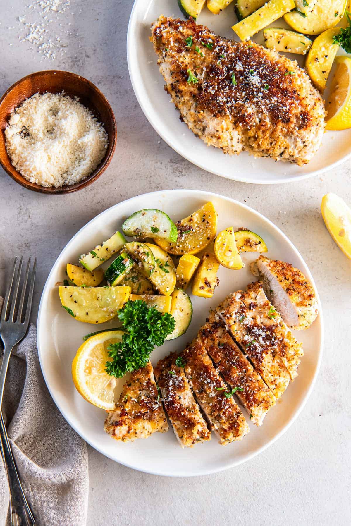 Breaded sliced chicken on a plate with zucchini next to a small bowl of Parmesan.
