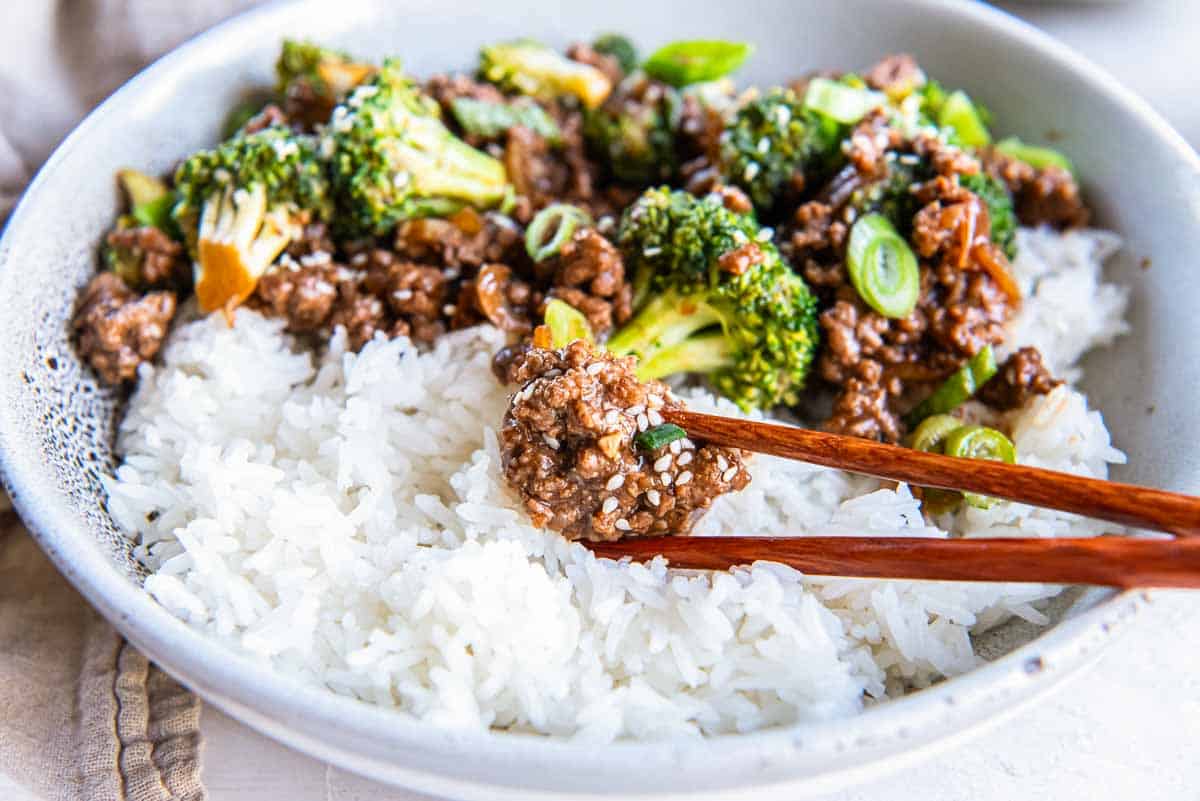 Chopsticks holding a piece of cooked ground beef over a bowl of rice.