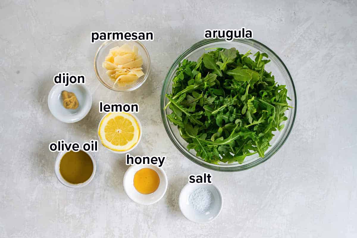 Baby arugula, parmesan, half a lemon and other ingredients in bowls with text.