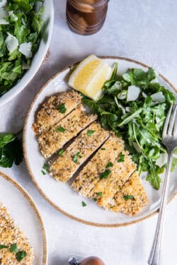 A sliced breaded chicken cutlet on a plate with arugula salad and a lemon wedge.