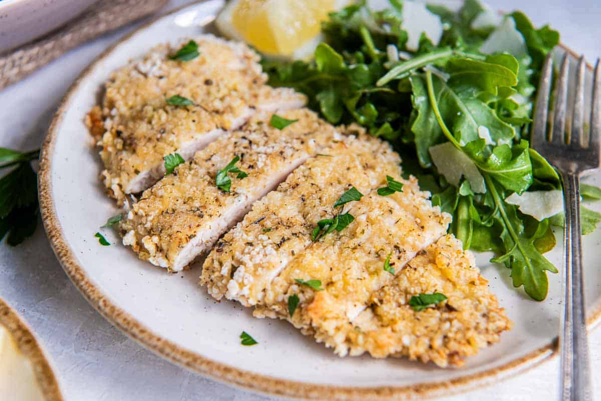 A sliced breaded chicken cutlet on a plate with arugula salad and a lemon wedge.