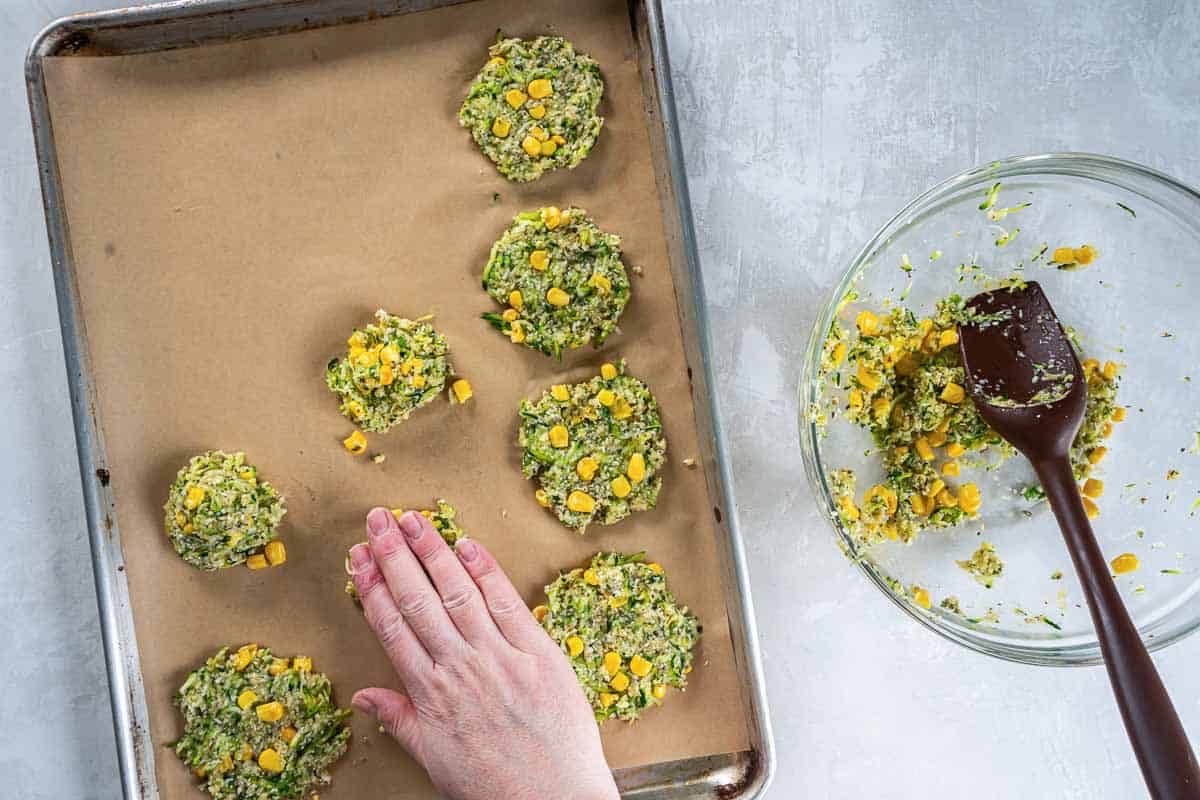 A hand patting down a zucchini corn fritter on a parchment paper lined baking sheet.