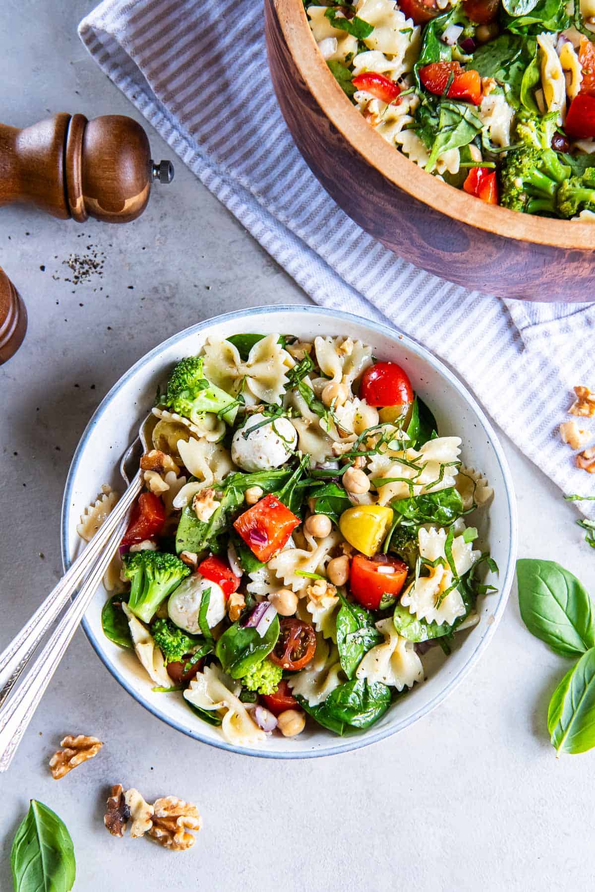 Spinach pasta salad in a small white bowl with forks next to a wood serving bowl.