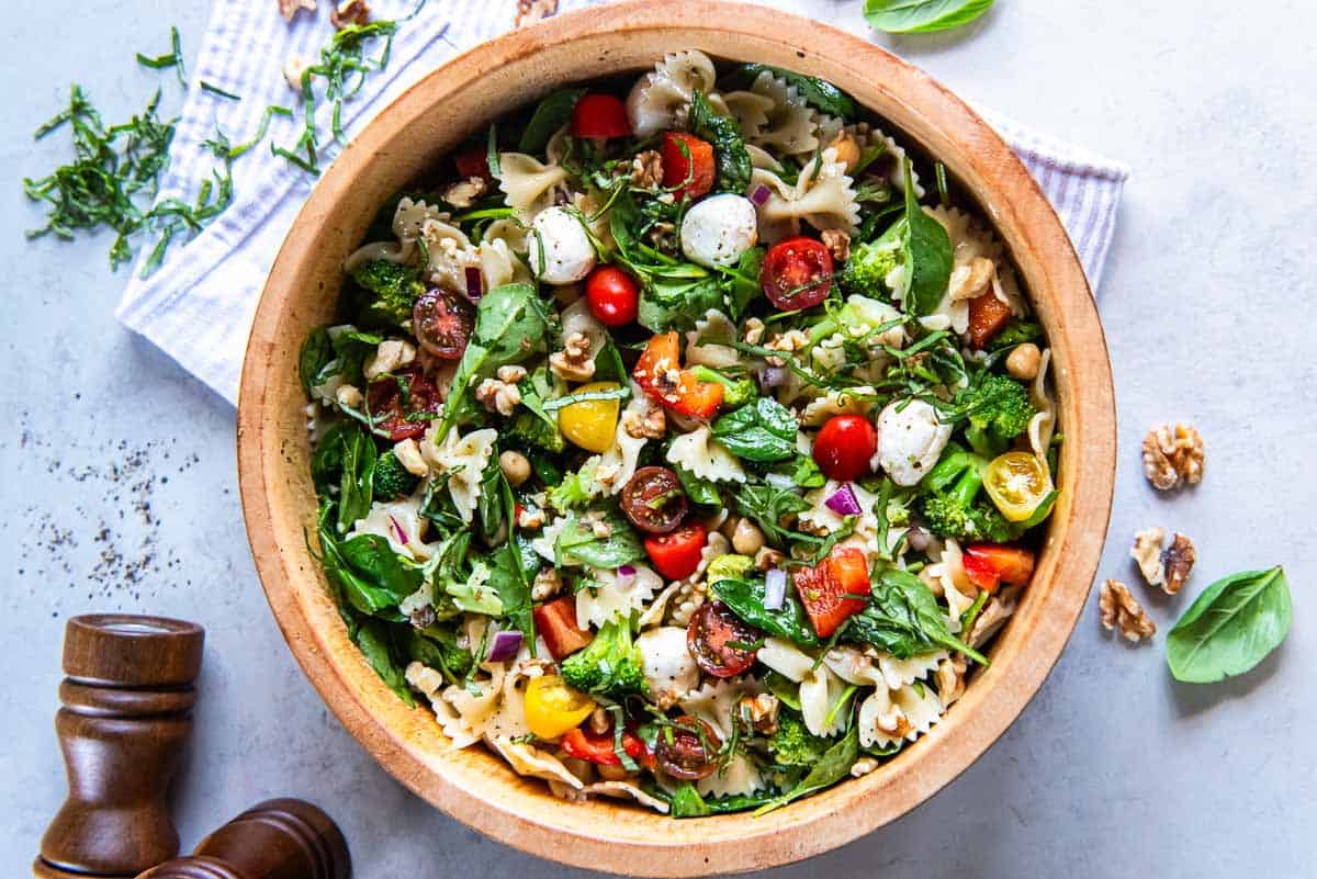 A colorful pasta salad in a wooden bowl filled with bowtie pasta, cherry tomatoes, mozzarella balls, spinach, chickpeas, broccoli florets, red onions, and fresh basil. A pepper grinder and loose basil leaves are nearby on a light surface.