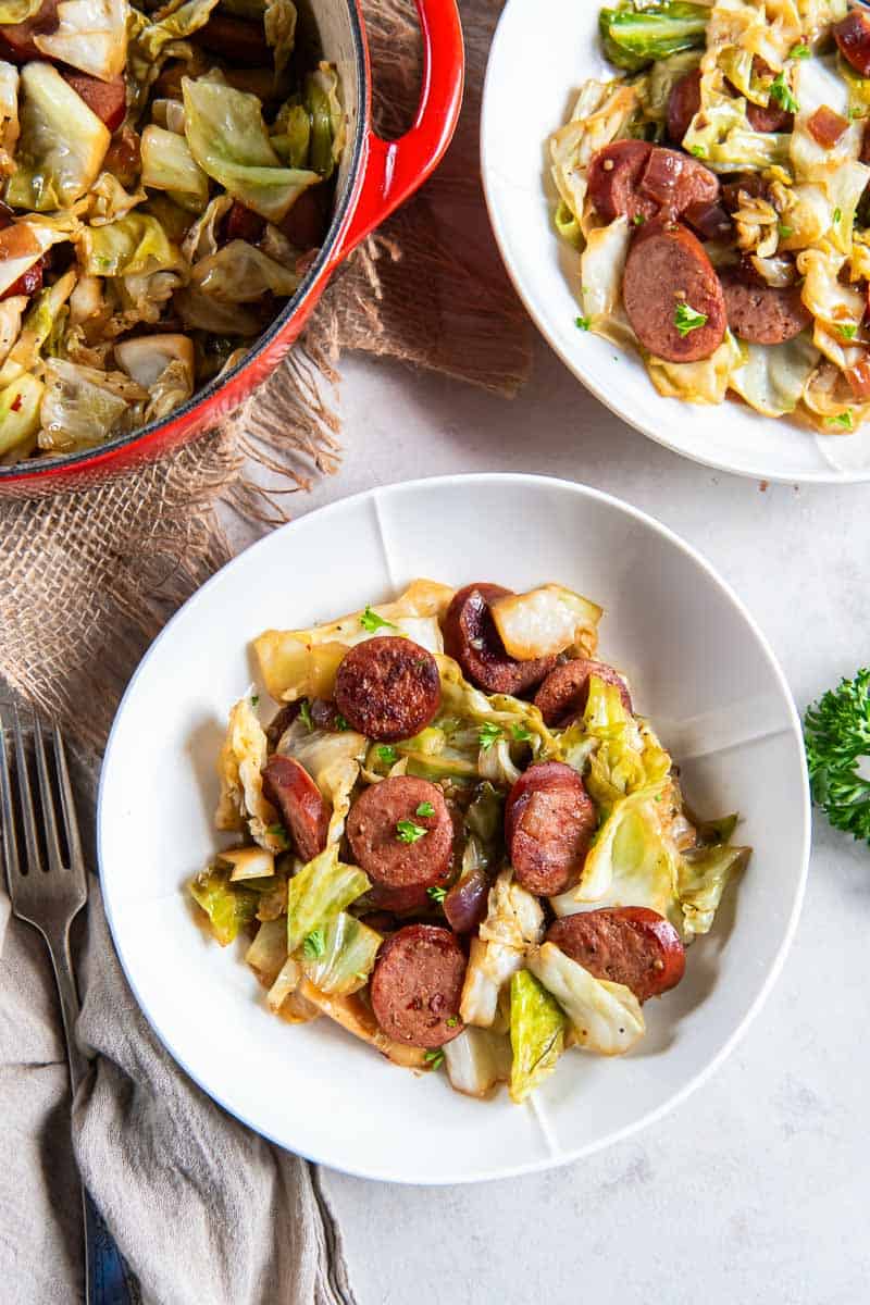 Two bowls of cabbage and sausage next to a pot.
