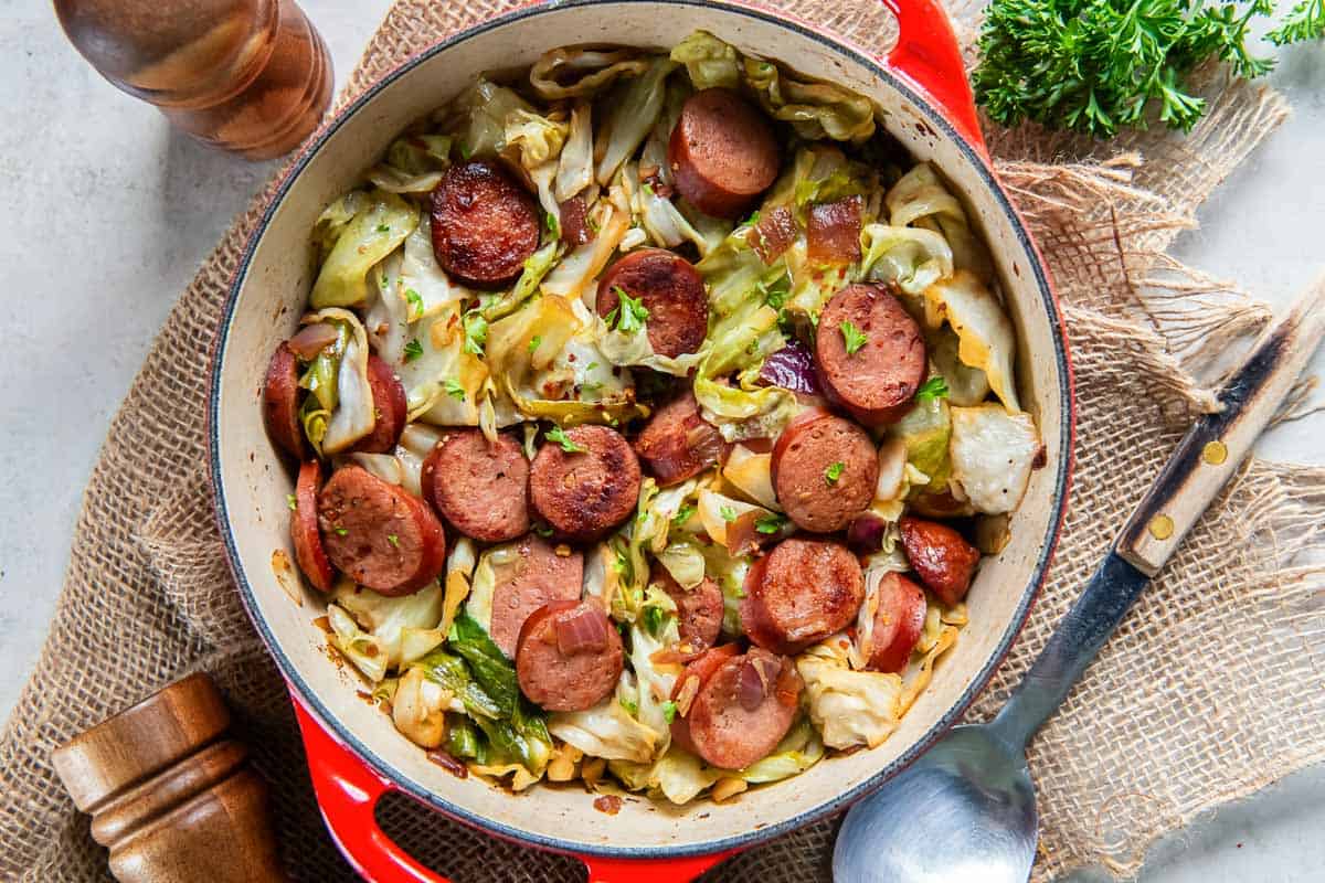 A spoon lying next to a pan filled with cabbage and sausage.