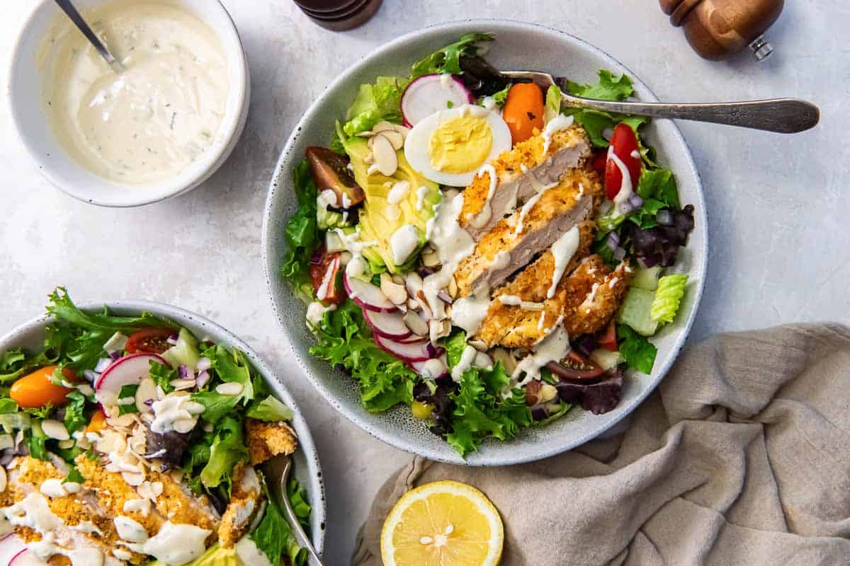 Two bowls of crispy chicken salad next to a small bowl of creamy salad dressing.