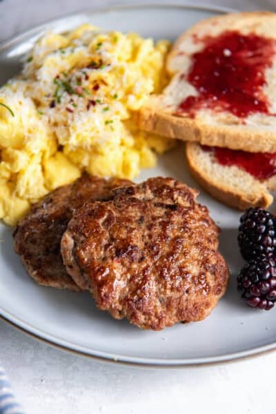 Two turkey breakfast sausage patties on a plate with scrambled eggs and toast with jelly.