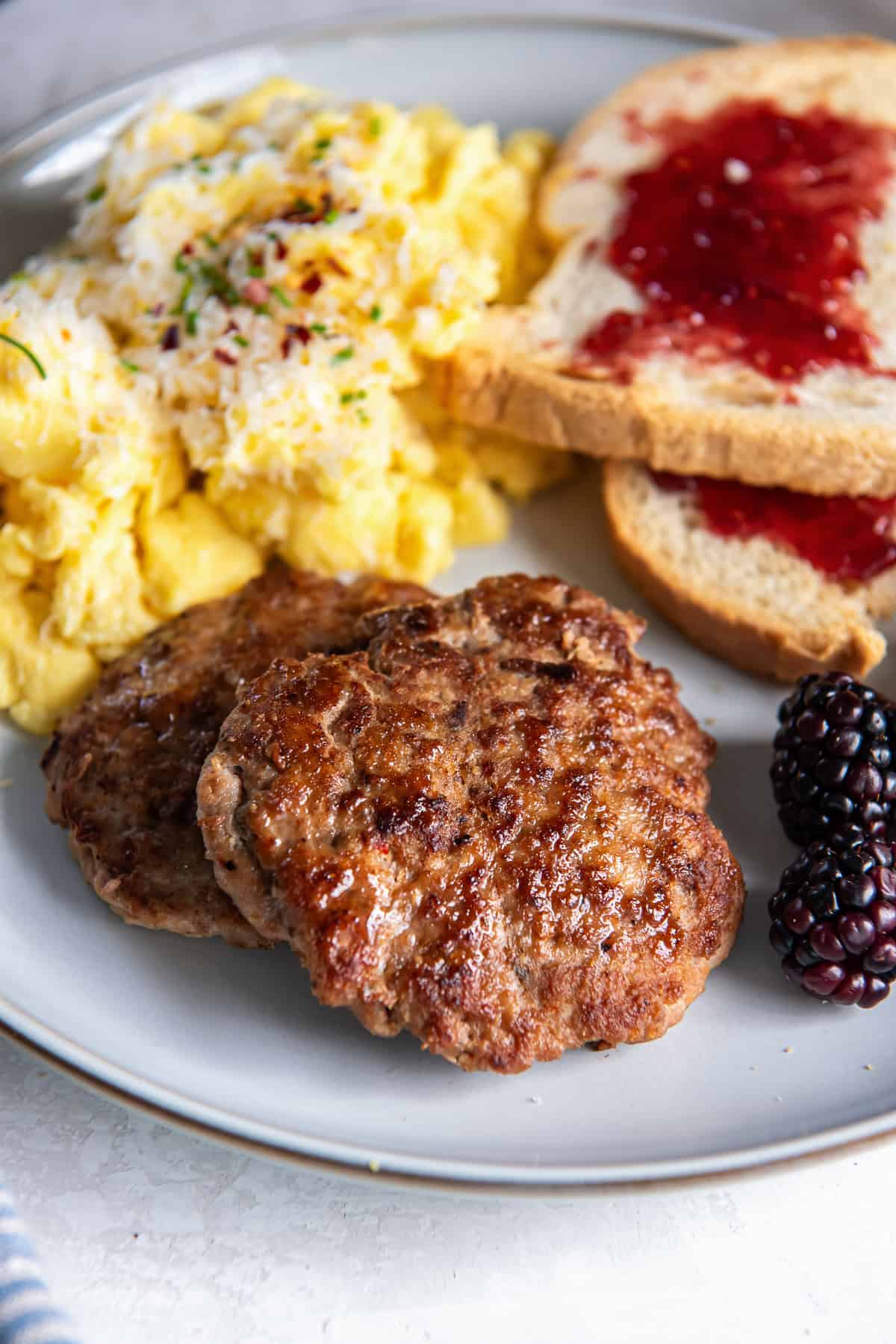 Two turkey breakfast sausage patties on a plate with scrambled eggs and toast with jelly.
