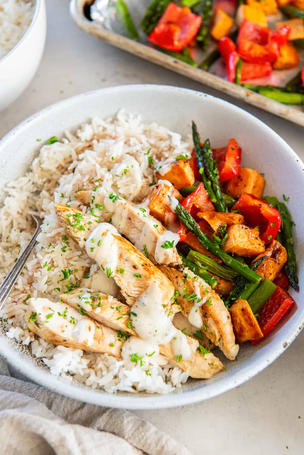 A chicken and roasted vegetable rice bowl next to a baking sheet filled with veggies.