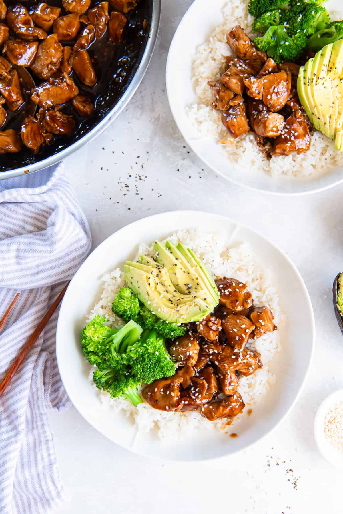 Two chicken teriyaki rice bowls with broccoli and avocado next to a skillet.