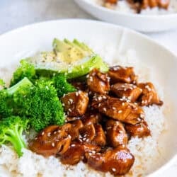 Bite size chicken teriyaki, rice, brocoli, and sliced avocado in a white bowl.