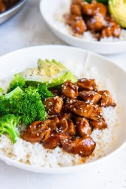 Bite size chicken teriyaki, rice, brocoli, and sliced avocado in a white bowl.