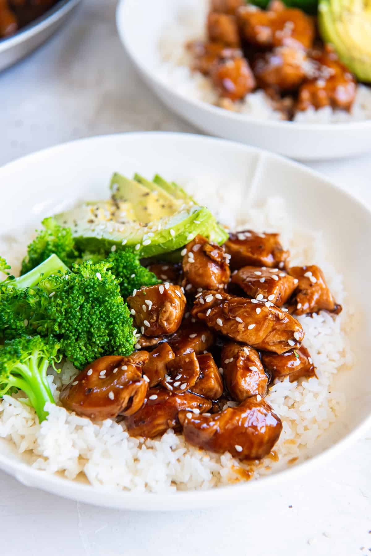 Bite size chicken teriyaki, rice, brocoli, and sliced avocado in a white bowl.