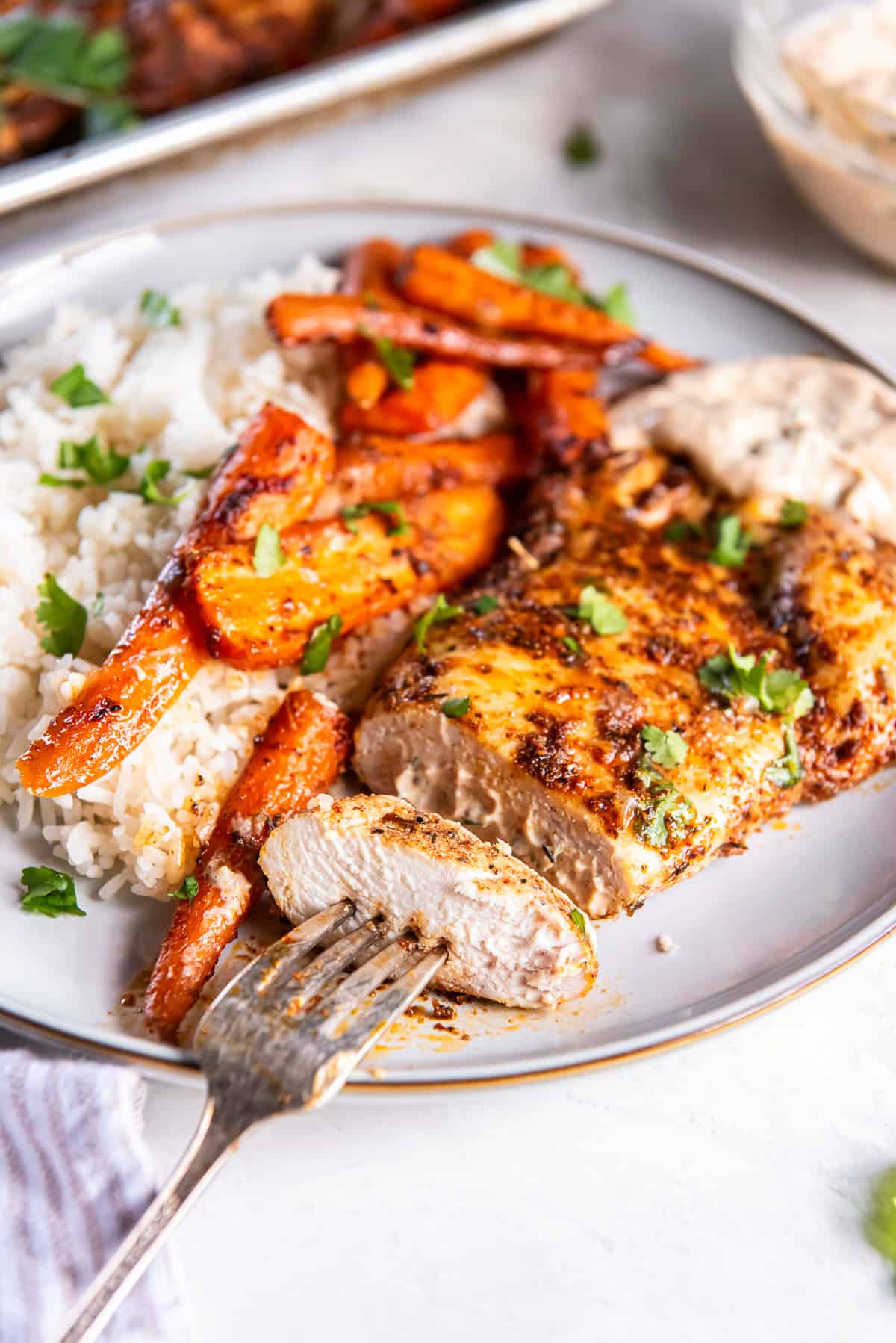 A fork piercing a slice of chicken resting on a plate with roasted carrots and rice.