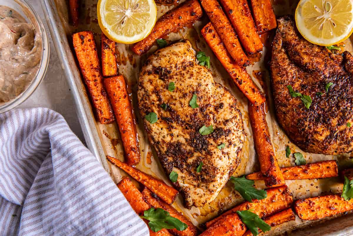 A sheet pan filled with cooked chicken breasts, roasted carrots and a halved lemon.