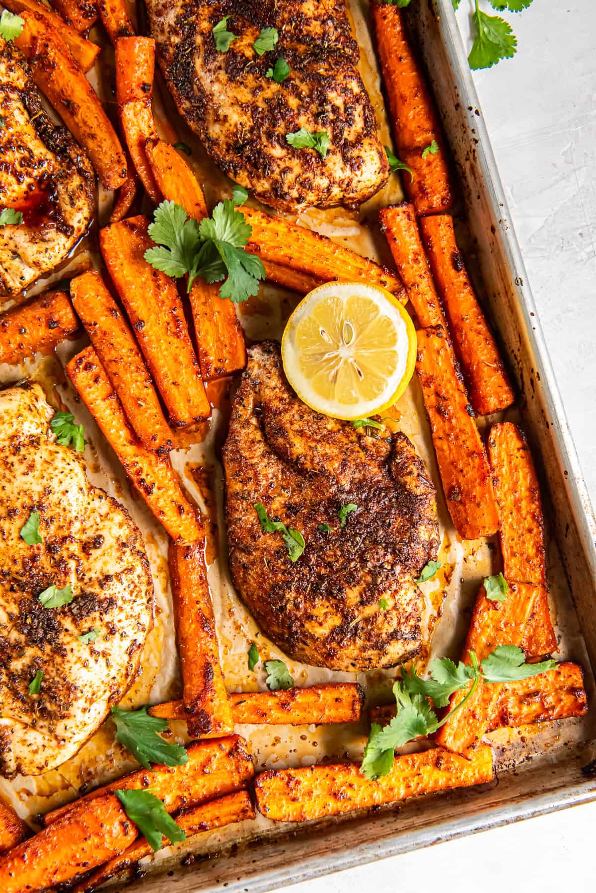 Seasoned cooked chicken breasts on a baking sheet with roasted carrots and lemon halves.