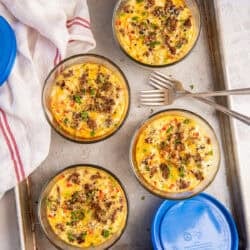 Egg bakes on a baking sheet in small glass containers topped with sausage with blue lids lying next to them.