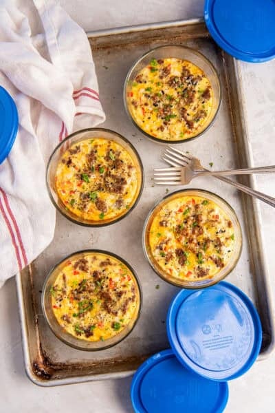 Egg bakes on a baking sheet in small glass containers topped with sausage with blue lids lying next to them.