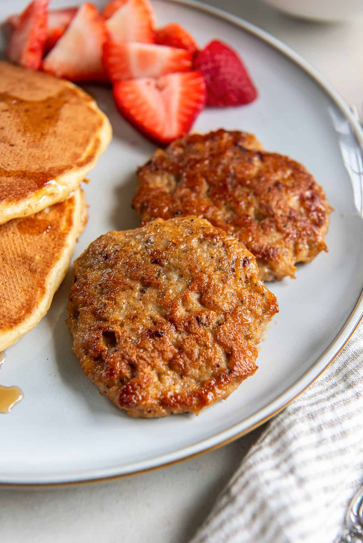 Chicken breakfast sausage patties, pancakes, and strawberries on a white plate next to a white cloth napkin.