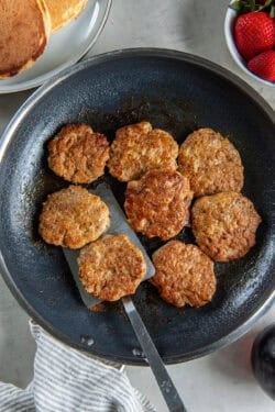 A spatula resting under chicken breakfast sausage in a skillet surrounded by a plate of pancakes and strawberries.