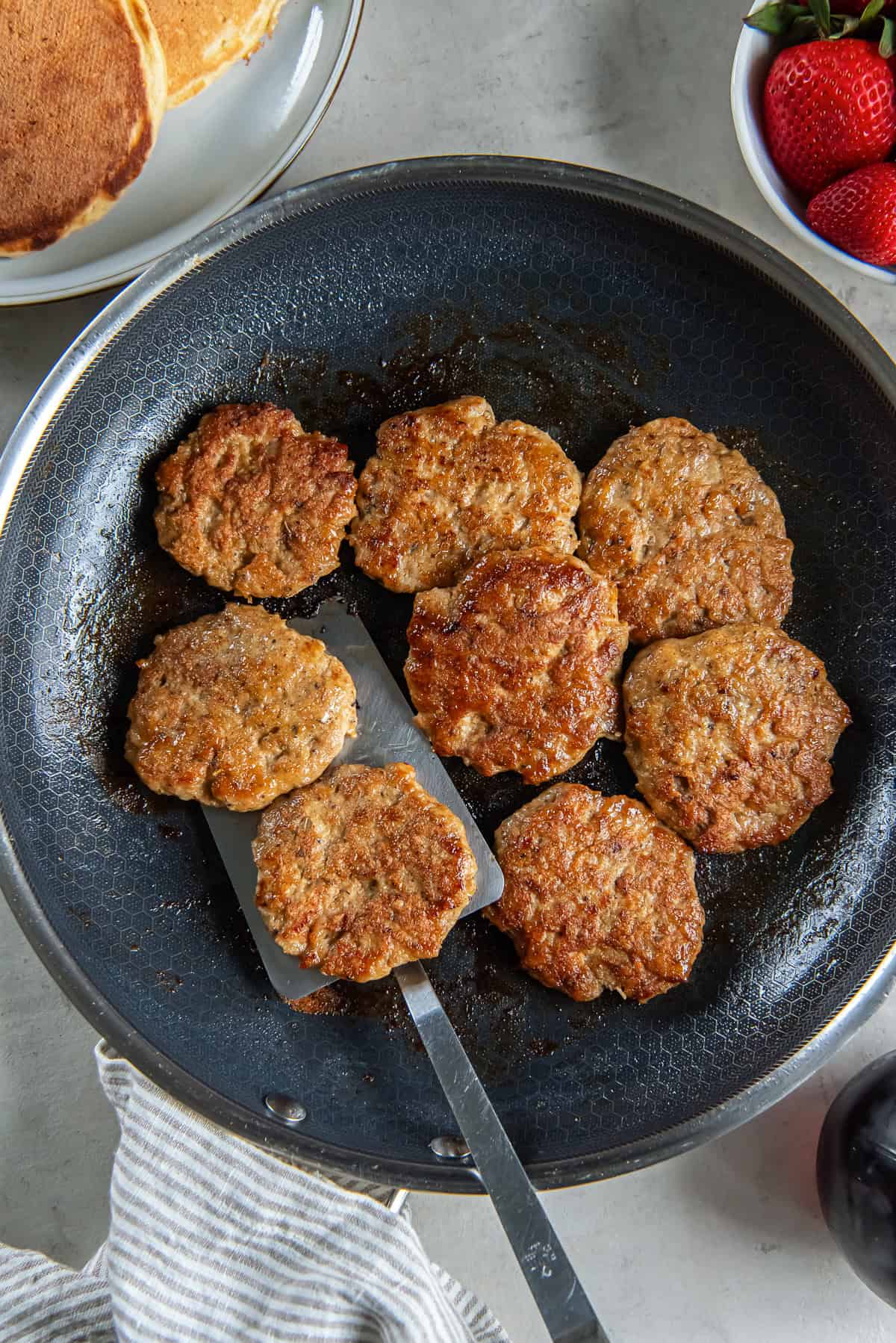 A spatula resting under chicken breakfast sausage in a skillet surrounded by a plate of pancakes and strawberries.