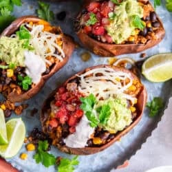 Four sweet potates stuffed with turkey taco filling with corn and black beans garnished with yogurt sauce, cheese, and guacamole on a metal platter.