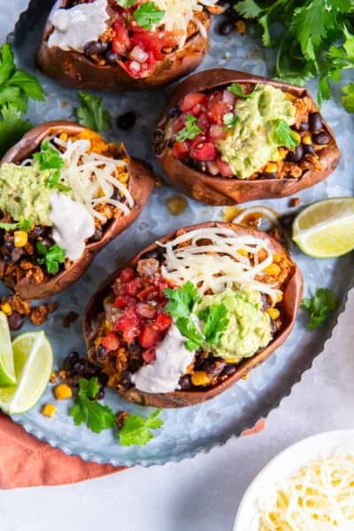 Four sweet potates stuffed with turkey taco filling with corn and black beans garnished with yogurt sauce, cheese, and guacamole on a metal platter.