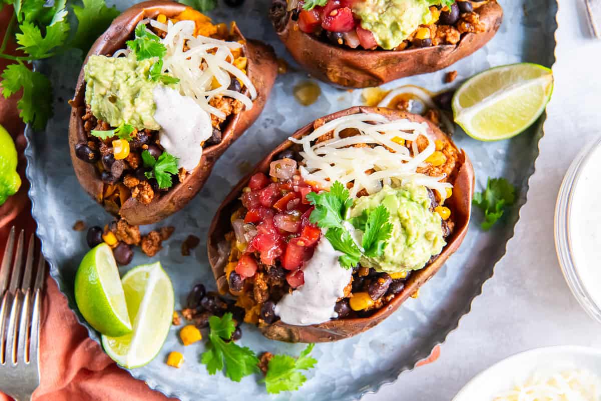 Taco stuffed sweet potatoes with salsa, yogurt sauce, cheese, and guacamole on a metal platter.