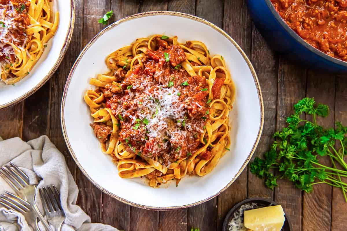 Bowl of tagliatelle with chicken bolognese, Parmesan, and parsley, with a Dutch oven and herbs in the background.