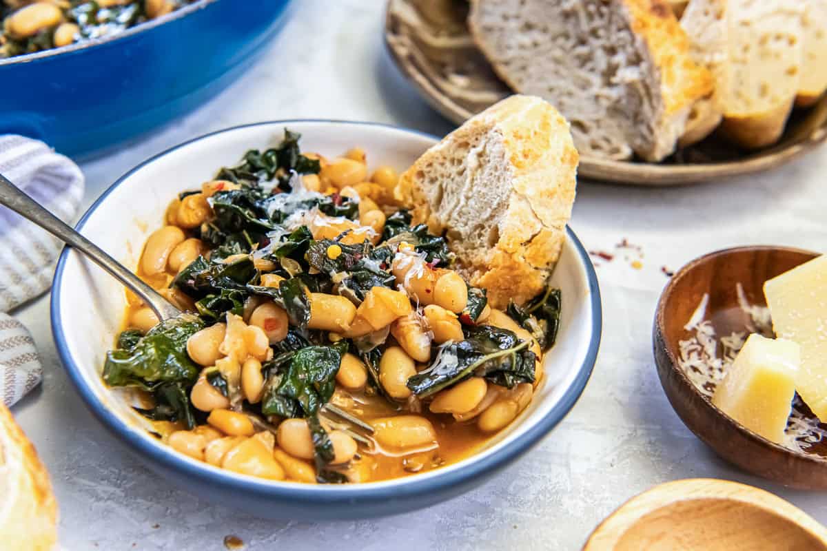 White beans and greens topped with Parmesan in a small bowl with a fork and a slice of French bread.