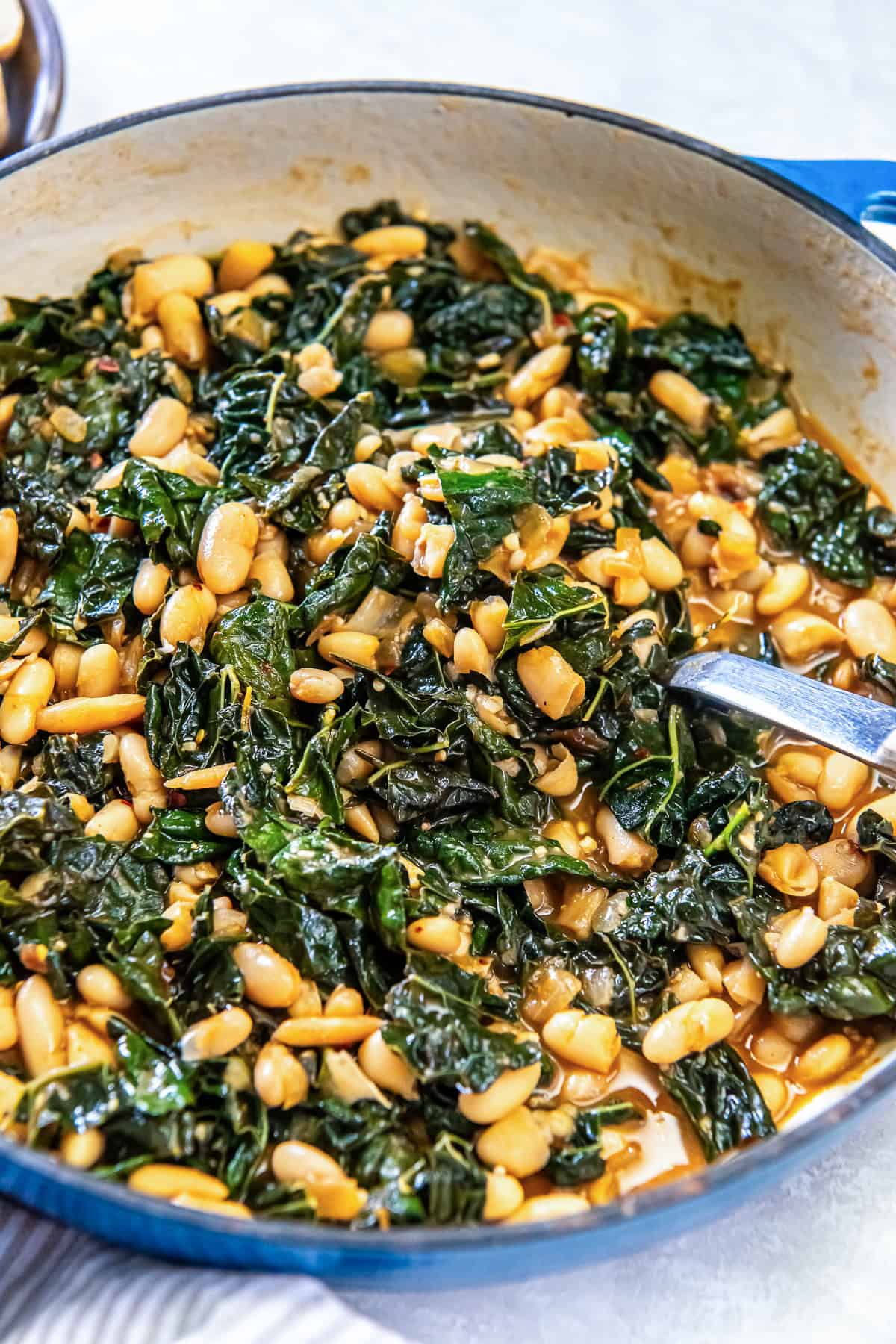 A spoon resing in a skillet with cannellini beans and kale in a braising liquid.