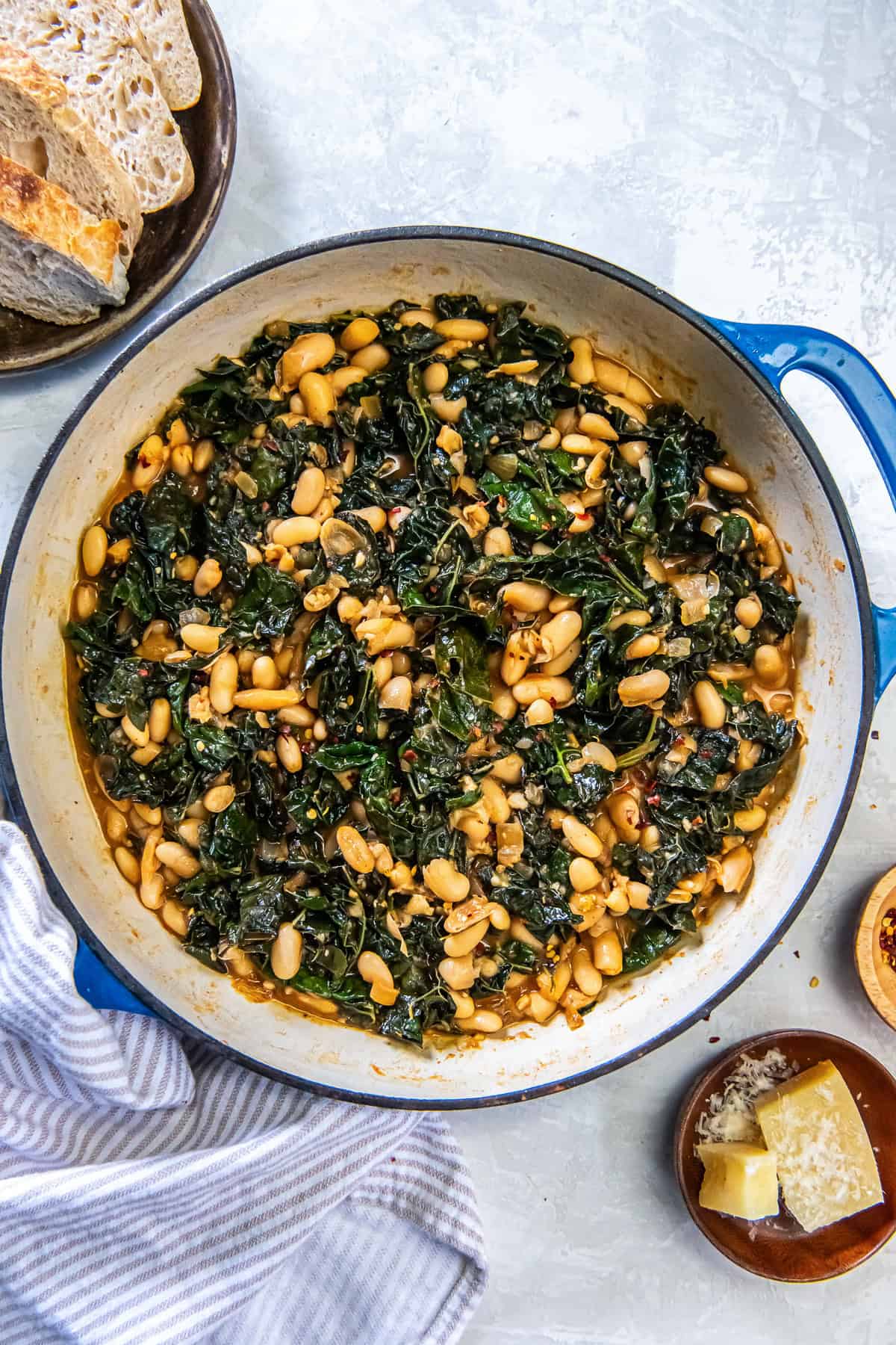 White beans and kale in a skillet next to a plate of bread and small bowl of Parmesan cheese.