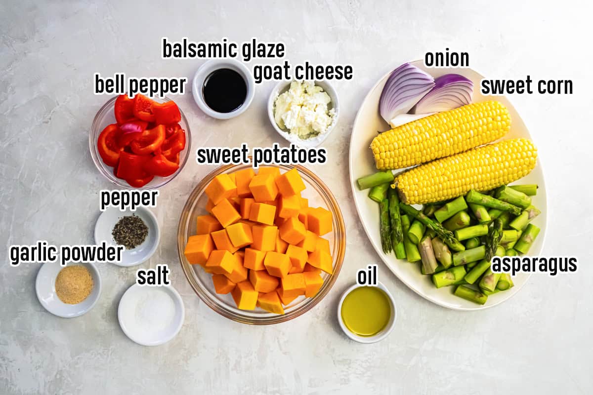 The ingredients for a roasted fall harvest salad, including sweet potatoes, bell pepper, asparagus, sweet corn, red onion, goat cheese, balsamic glaze, olive oil, and seasonings