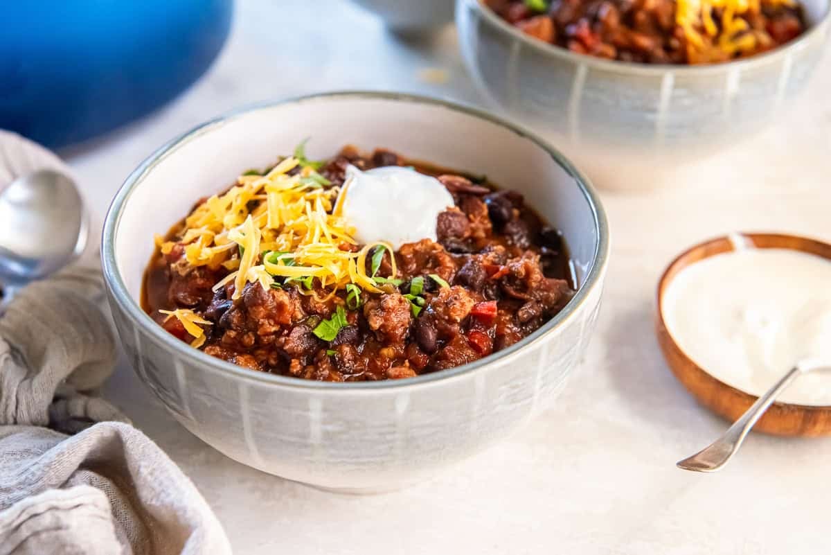 Bowl of turkey black bean chili topped with shredded cheddar, sour cream, and chopped cilantro, served with a spoon and napkin on a light surface.