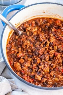 A spoon resting in a pot of turkey black bean chili with rich red sauce and hearty mix of beans, turkey, and vegetables.