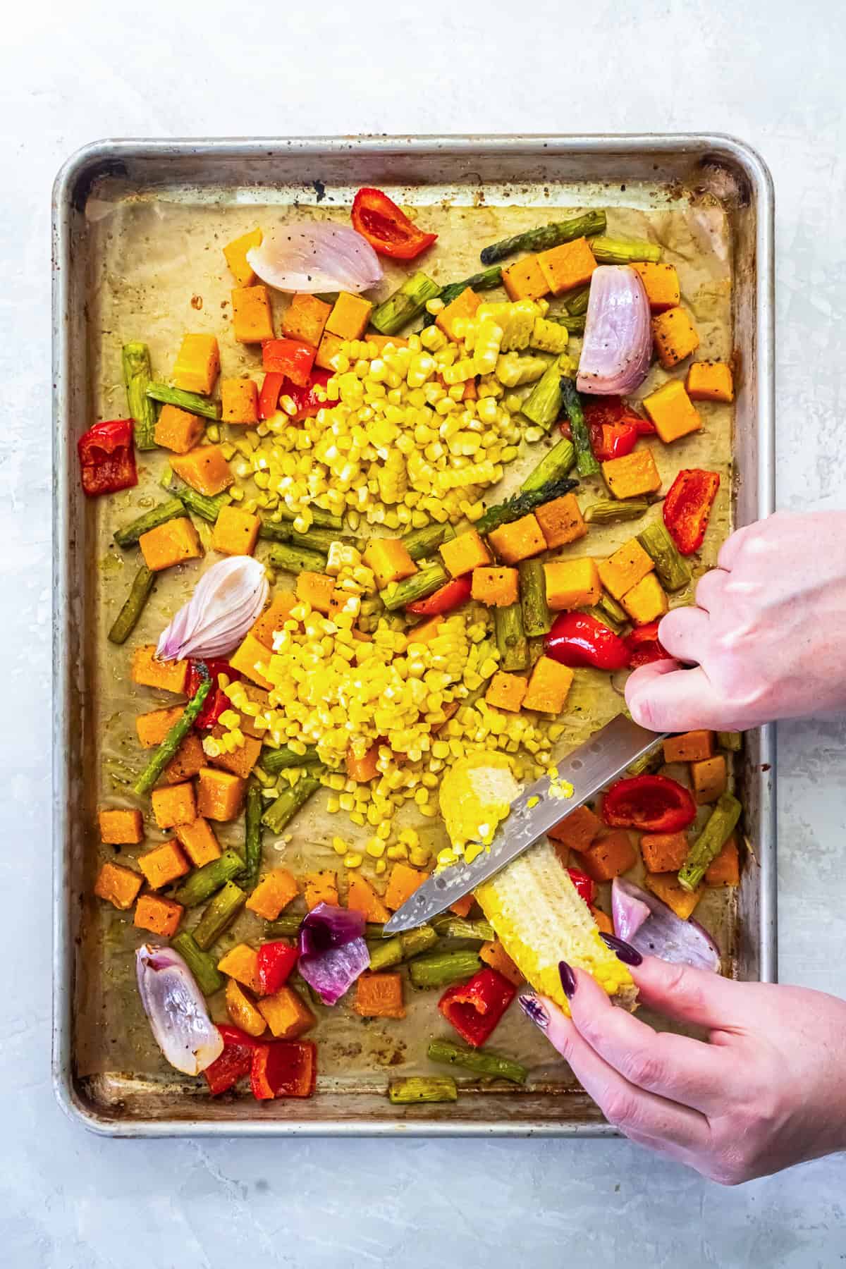 Hands slicing kernels off a roasted corn cob over a sheet pan filled with roasted sweet potatoes, asparagus, red bell pepper, and red onion.