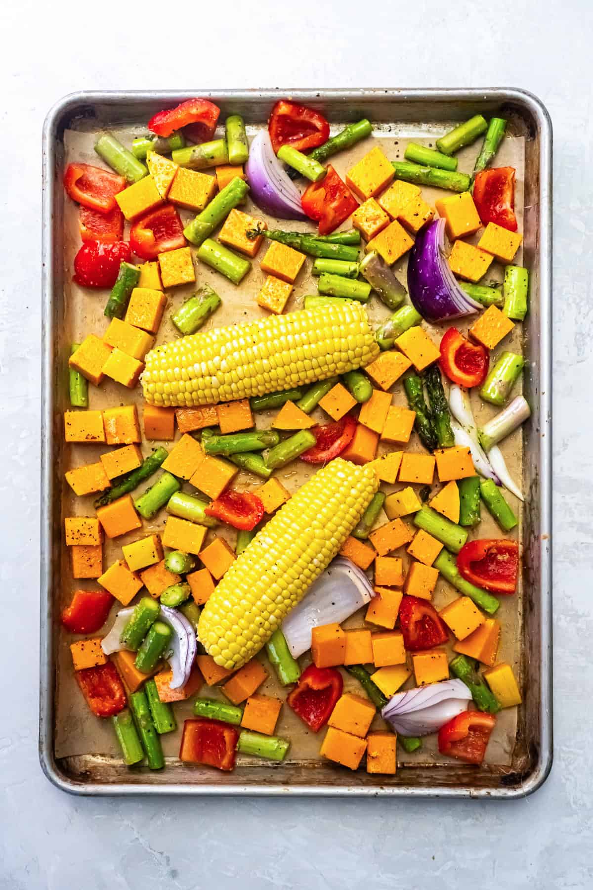 Sweet potatoes, asparagus, red bell pepper, and red onion arranged on a sheet pan with two ears of corn.