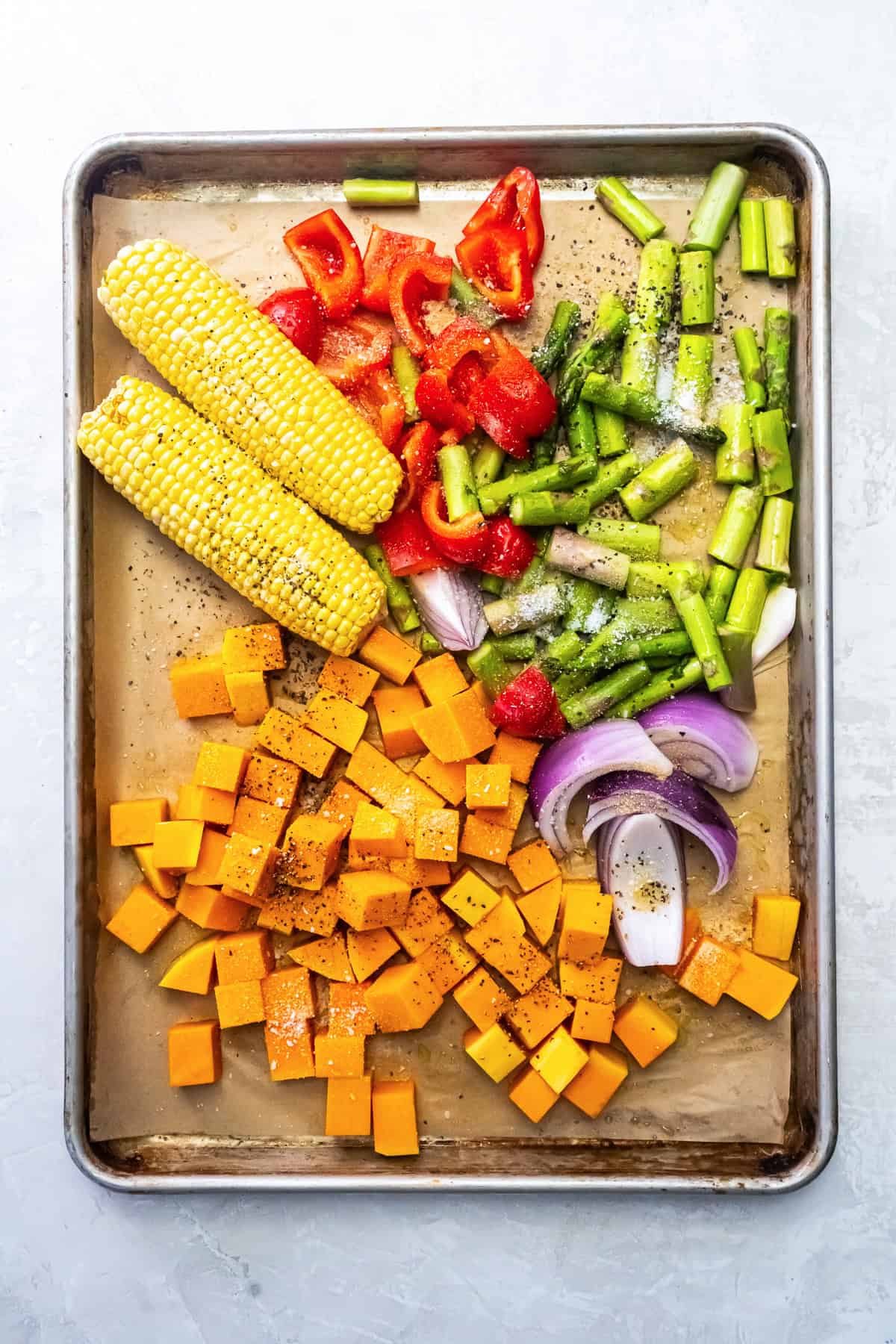 Sweet potatoes, asparagus, red bell pepper, red onion, and two ears of corn on a sheet pan, seasoned and ready to roast